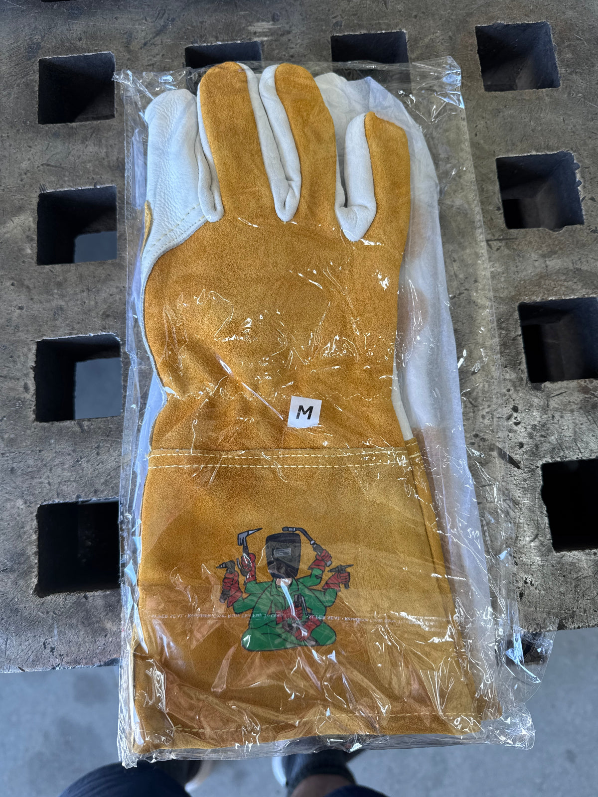 Pair of light brown gloves with white cuffs in clear plastic packaging on a concrete surface.